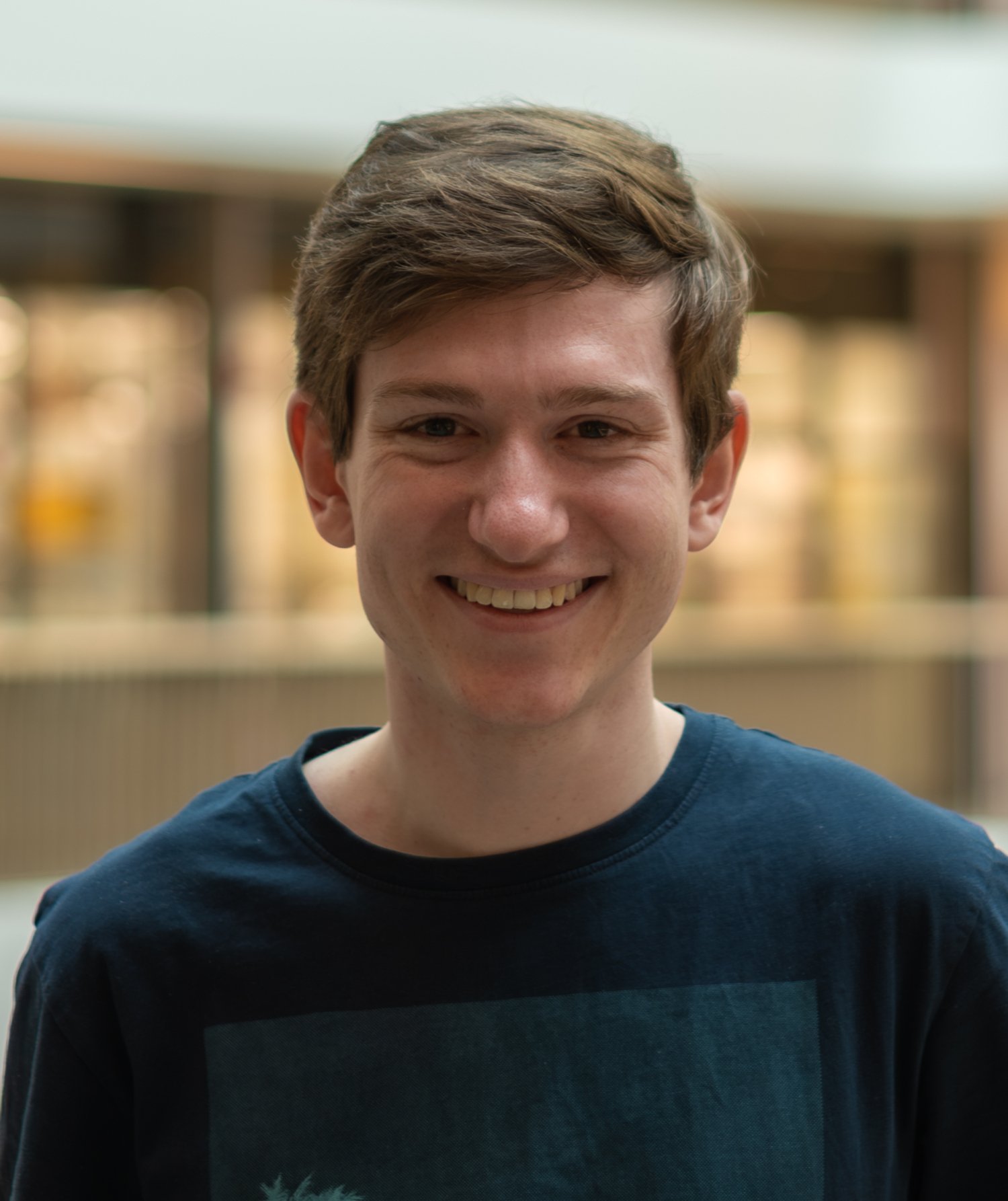 Person wearing a dark graphic T-shirt stands indoors in front of a blurred modern atrium background.