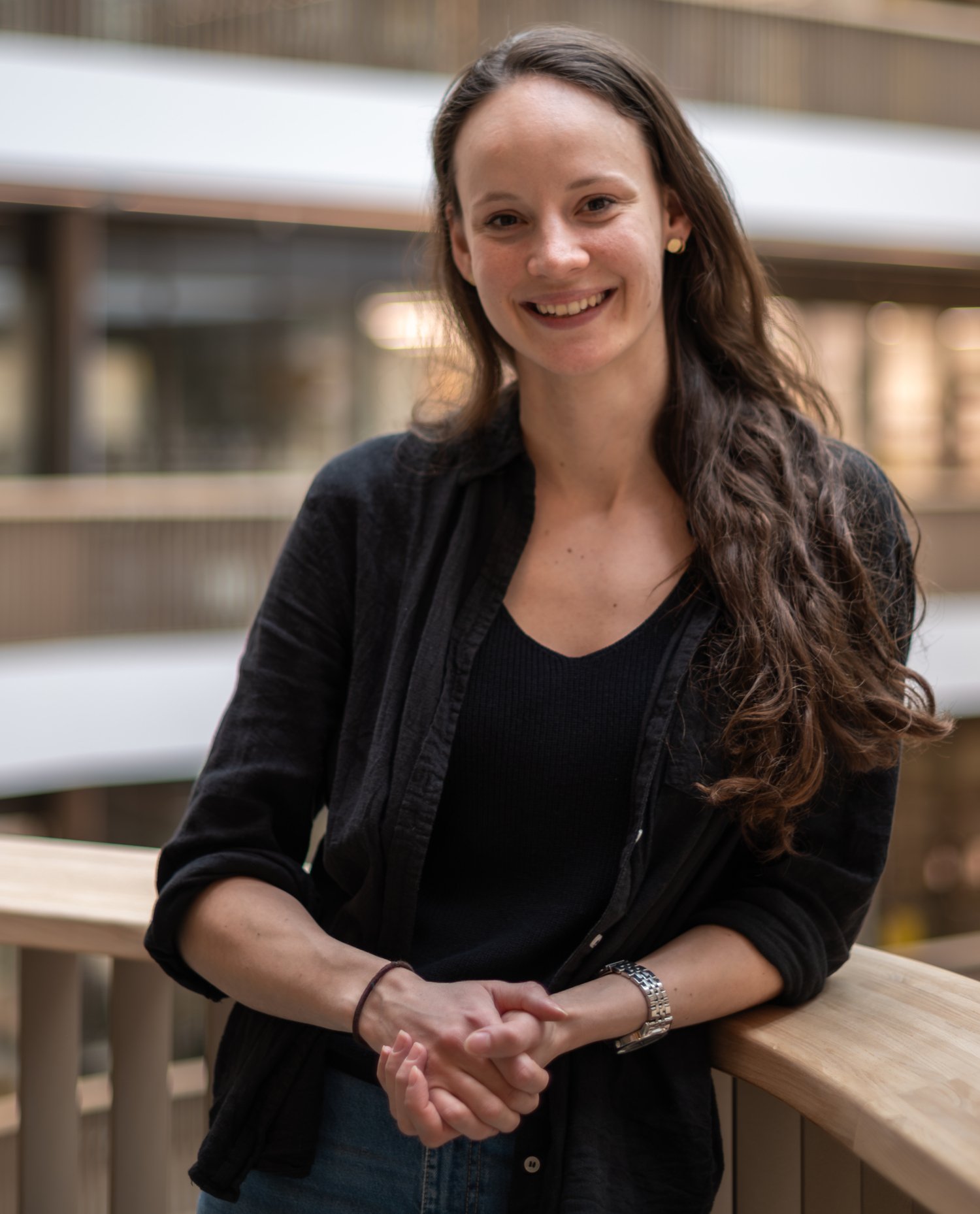 Smiling woman with long brown hair and a black shirt leaning against a railing.