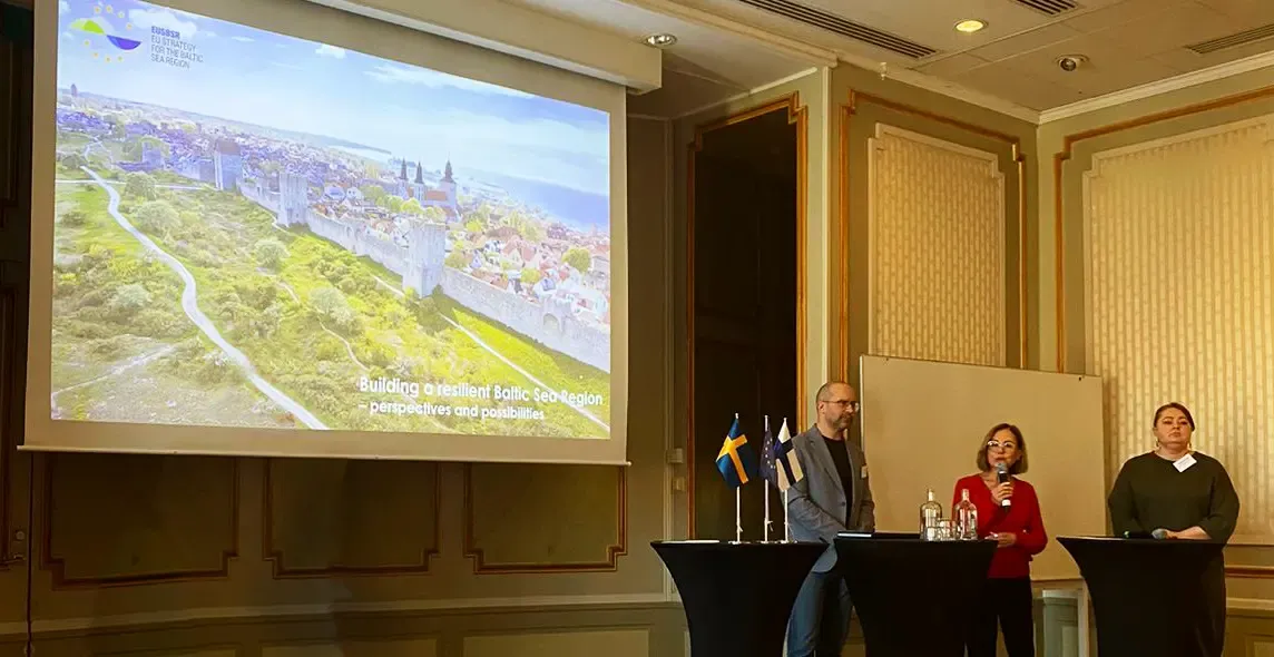 Three people, one of whom is speaking into a microphone, standing at a podium with three flags: Swedish, Finnish, and EU.