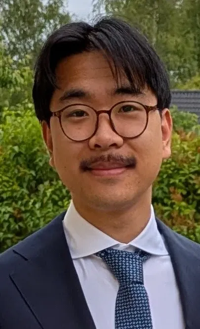 Dark‑haired man with glasses and a mustache, dressed in a blue blazer, white shirt, and blue checkered tie.