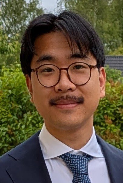 Dark‑haired man with glasses and a mustache, dressed in a blue blazer, white shirt, and blue checkered tie.