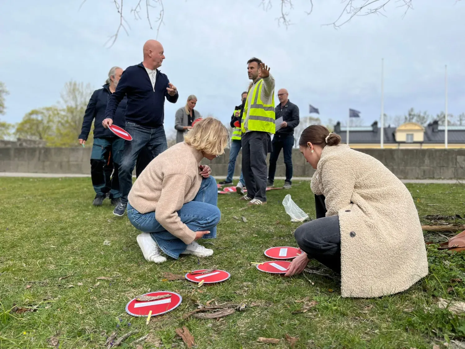 A group of people are standing in a grassy patch, looking at small round discs. One person is wearing a high-visibility vest.