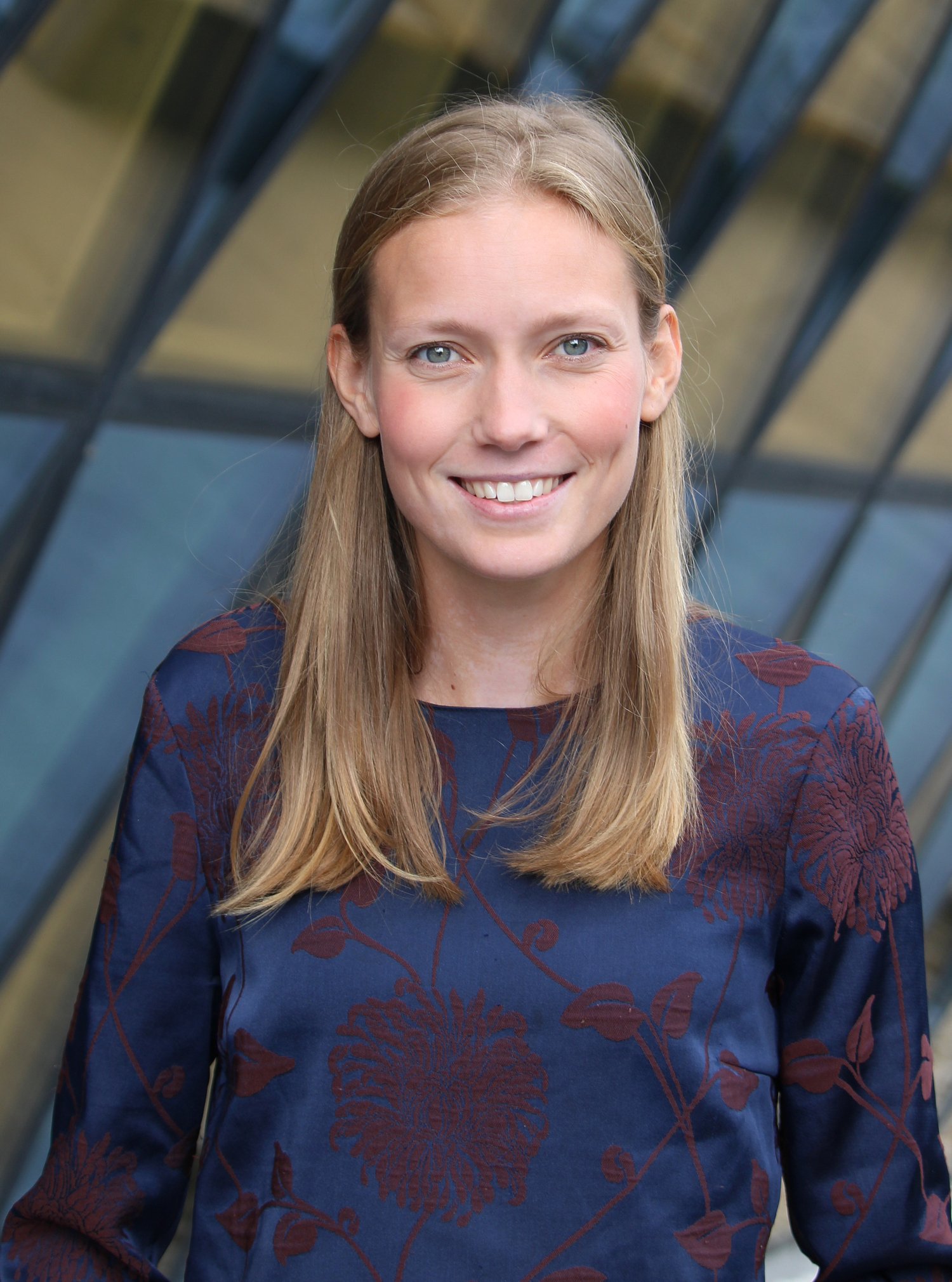 Woman with blonde hair wearing a blue top standing in front of a building with a glass front