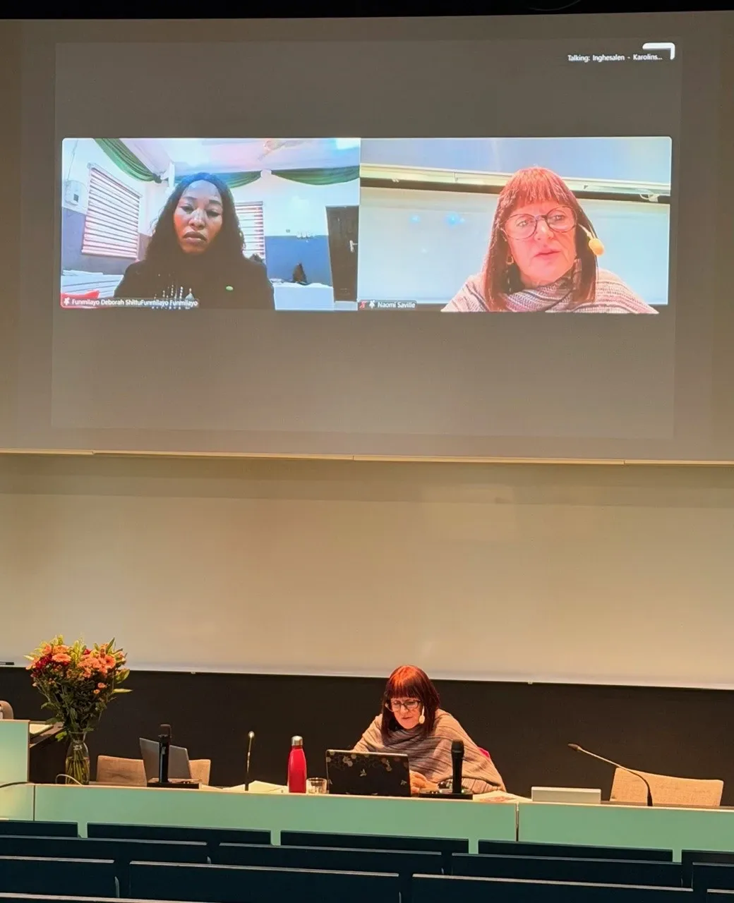 Two women during a Zoom meeting are shown on a large screen in a semiar room.