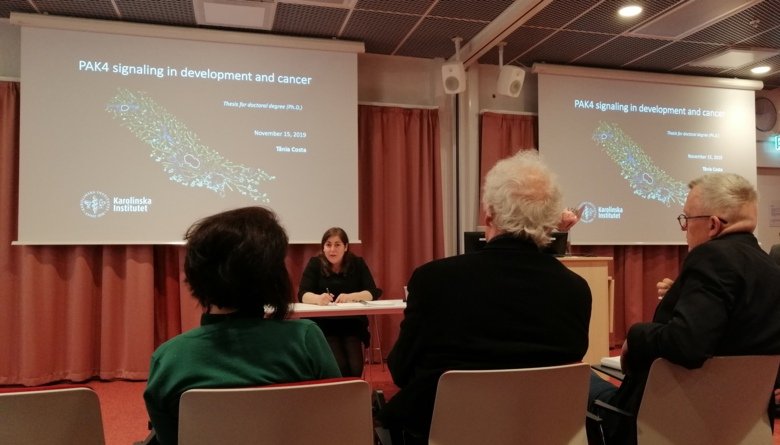 Woman presenting her thesis in front of the board, screens with her presentation is showing in the background