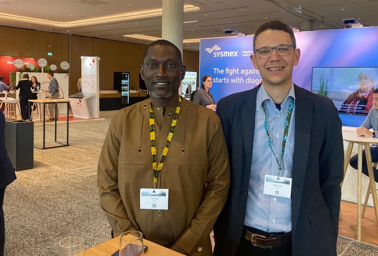 Two men standing beside eachother at a conference, smiling at the camera.