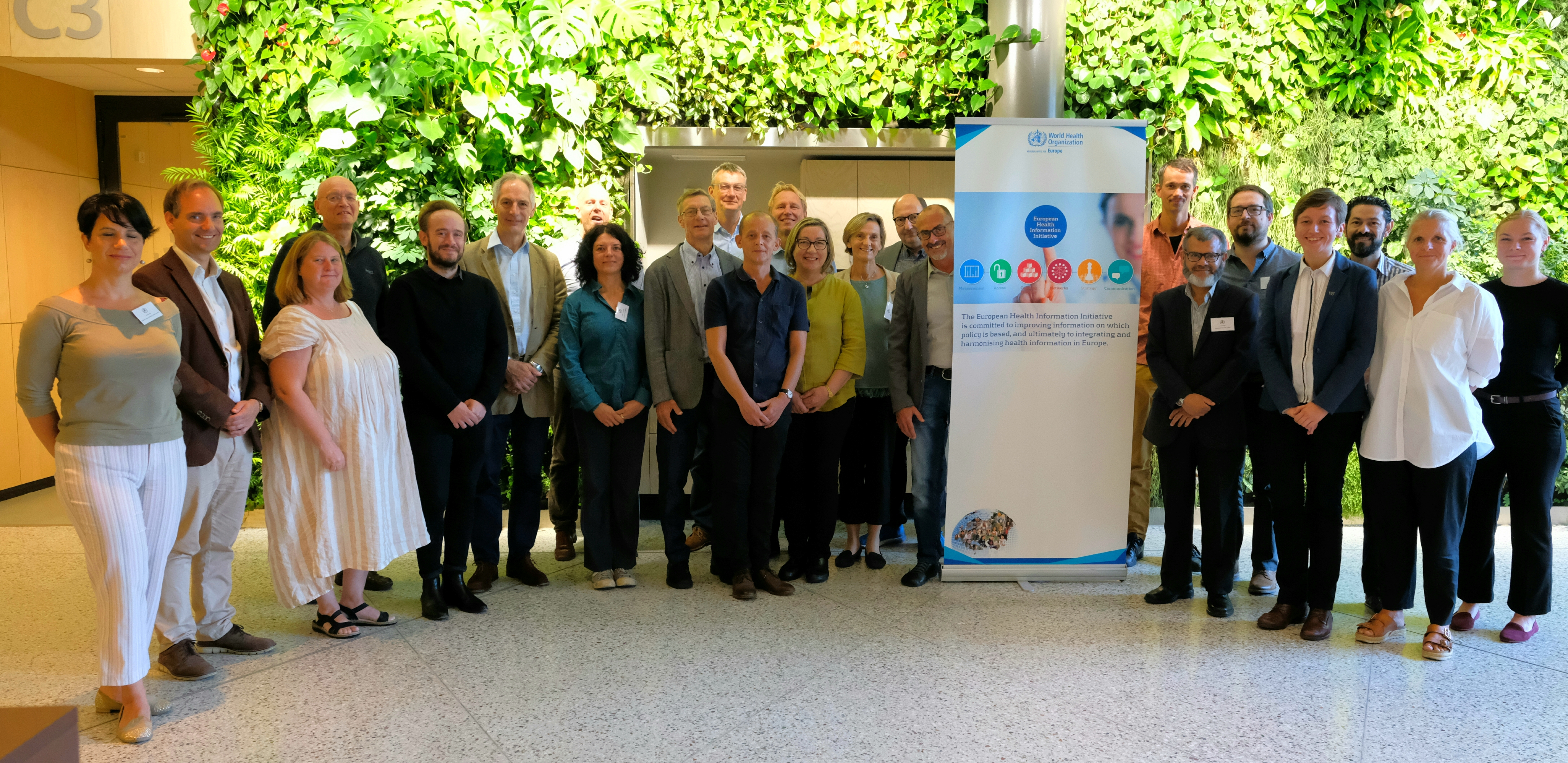 Group of people standing smiling in front of a wall of green plants.