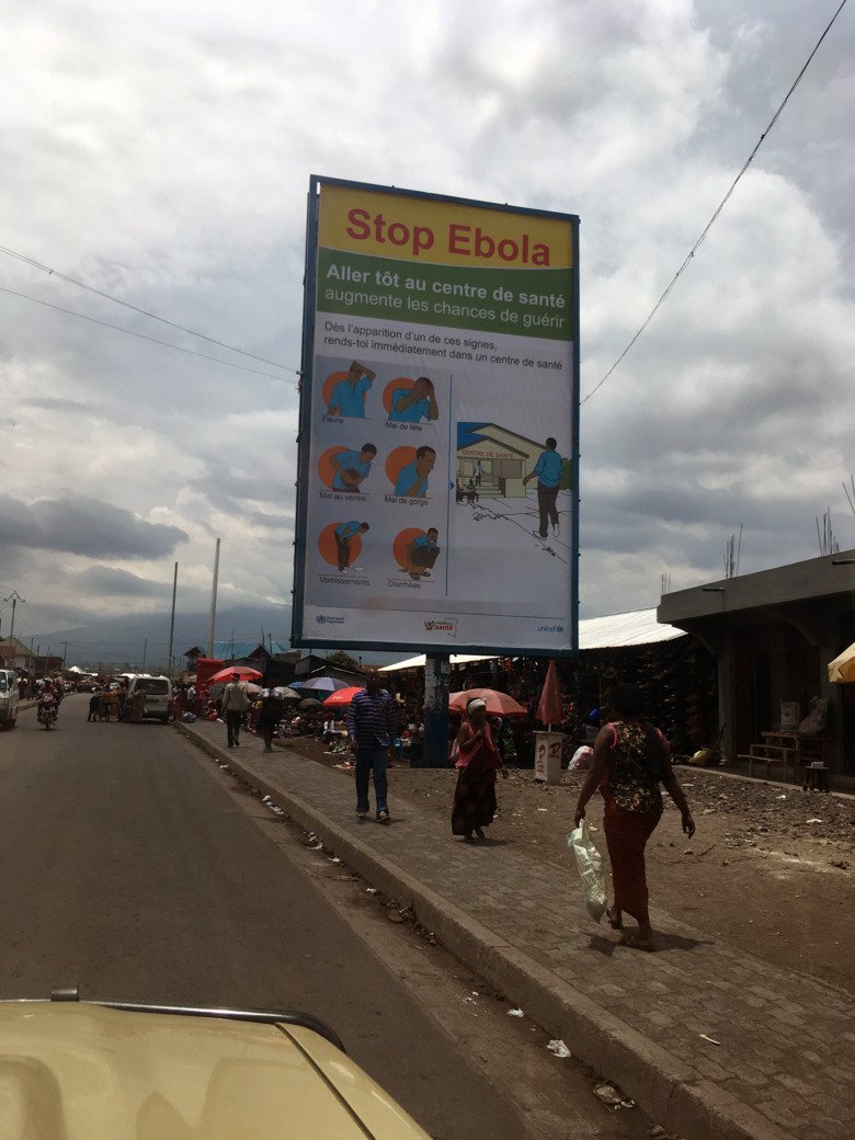 Sign with information about Ebola, along a street with cars, houses and people in the DR of Congo.