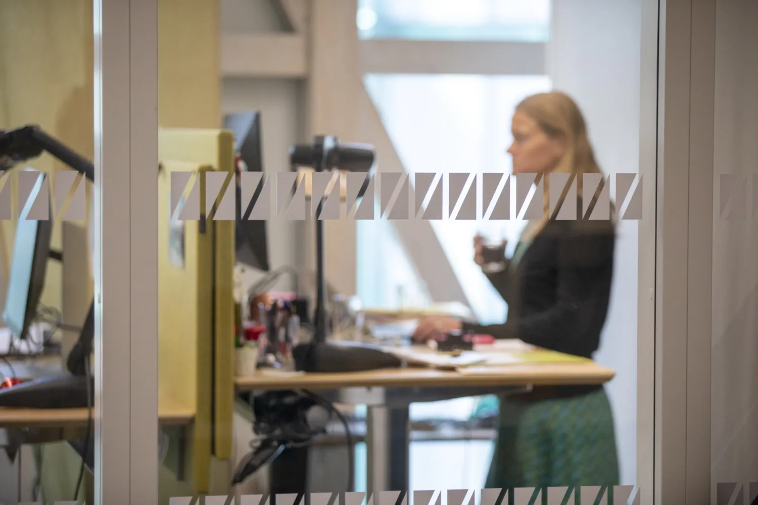 In the foreground: glass wall with motifs. In the background: Woman standing at a desk in open office landscape.