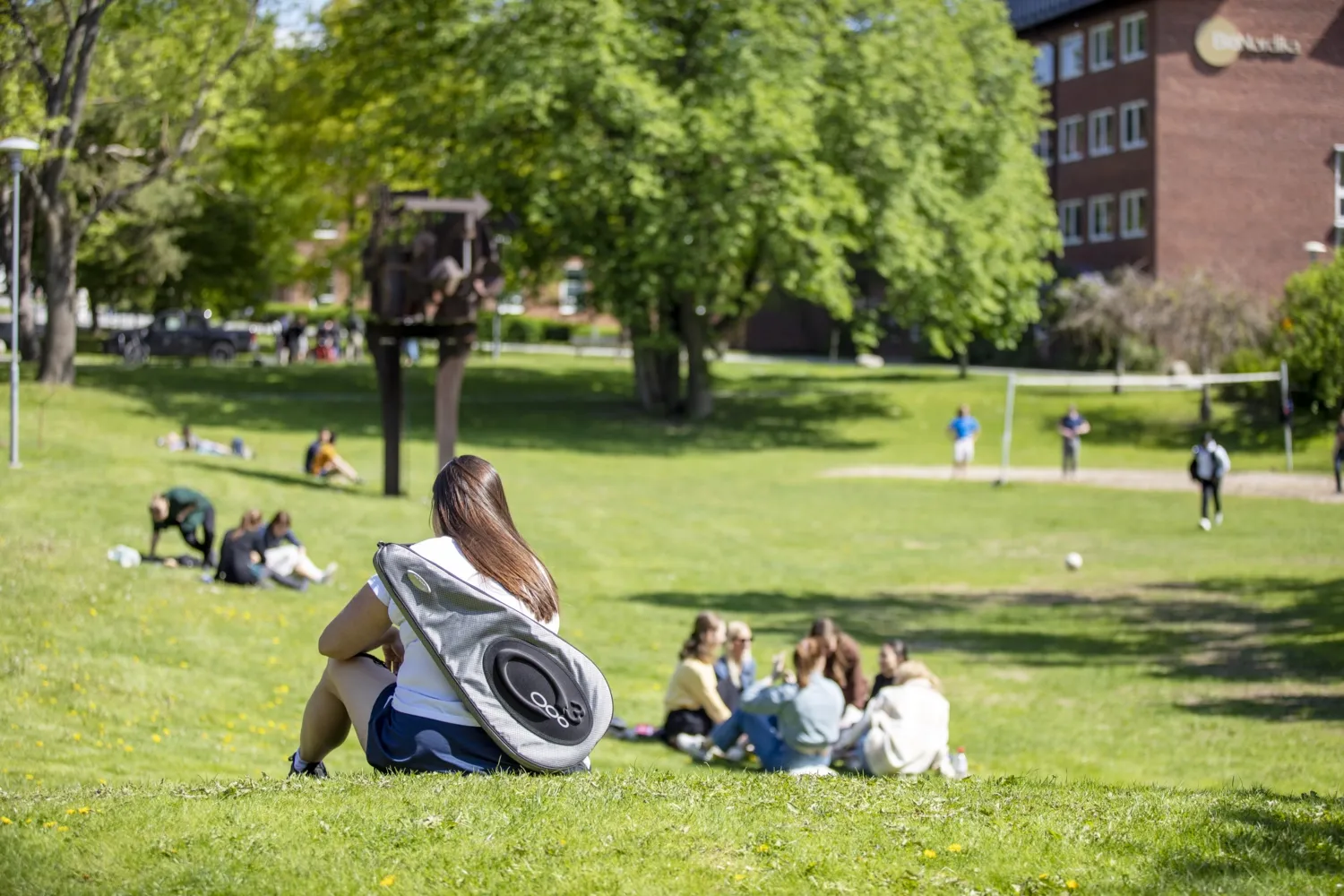 Woman with badminton racket bag sitting on her back on lawn on campus Solna, with people in the background.