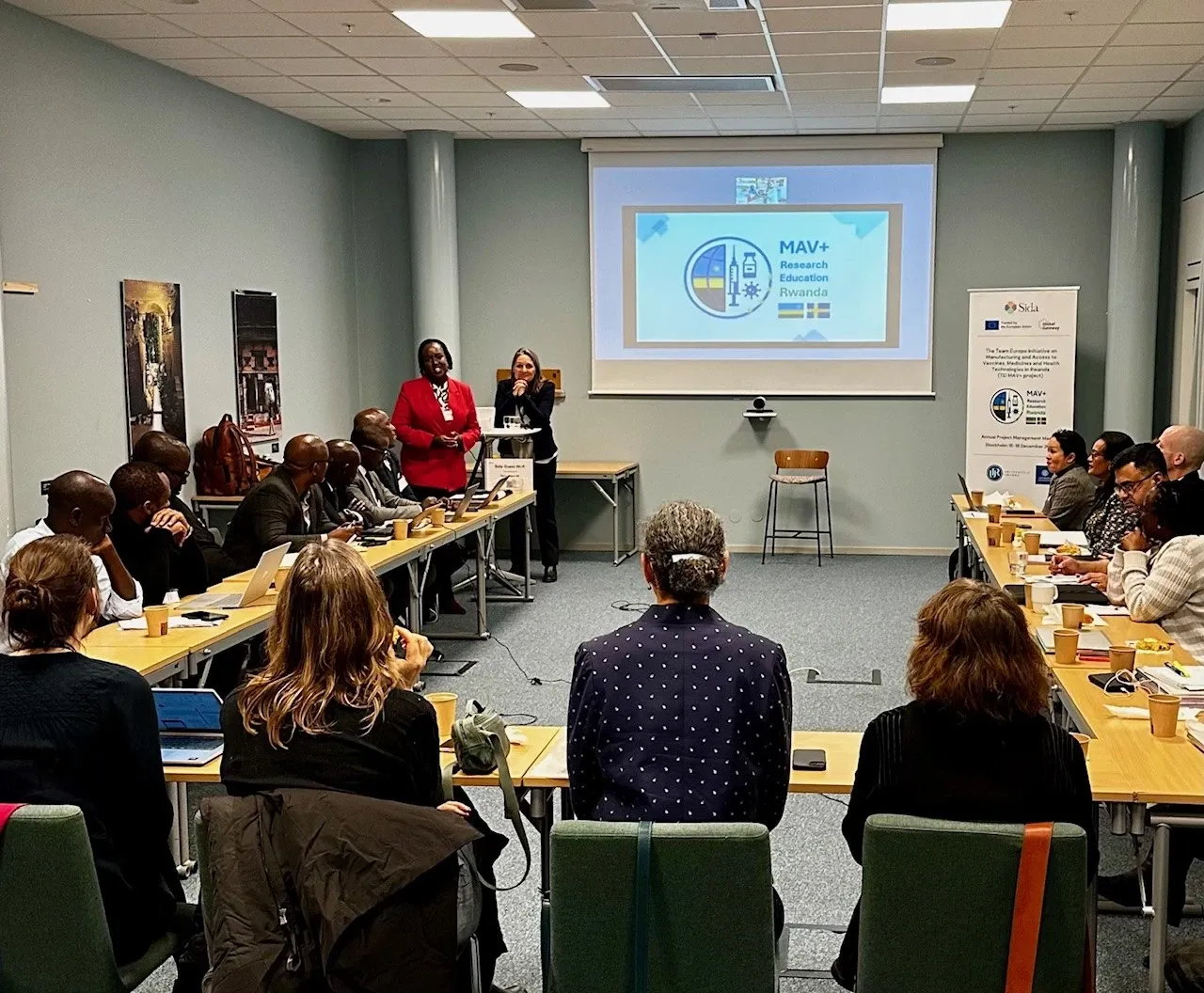 15 persons sitting around a u-shaped meeting table, listening to a woman standing at the front.