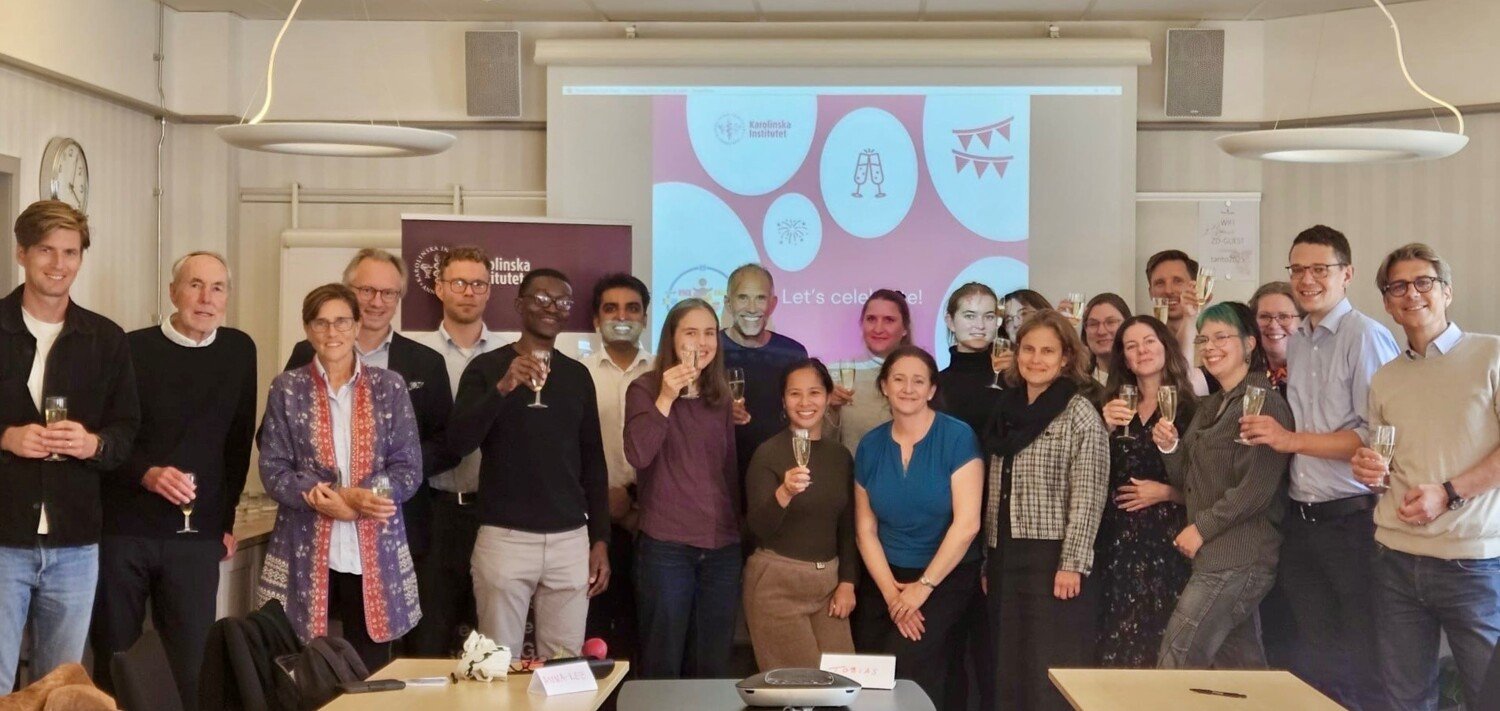 A group of peolpe are standing smiling in front of a sign saying Let's celebrate.