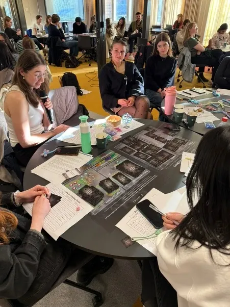 Students engage in a collaborative workshop session around round tables with cards on the table.