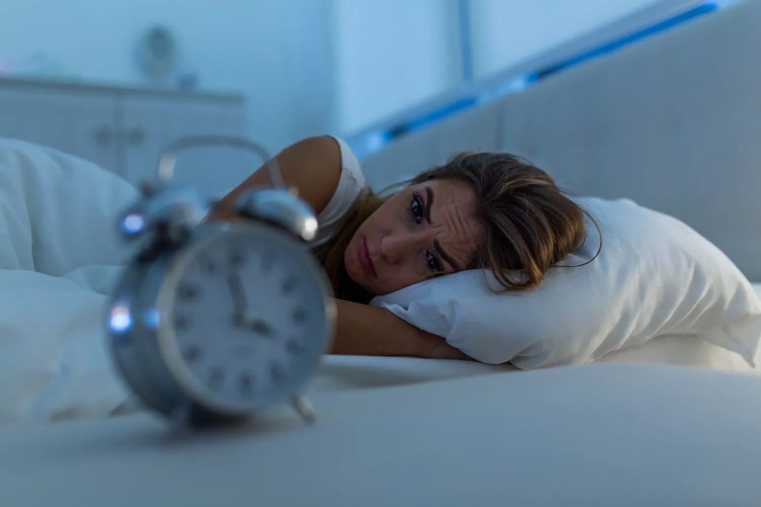 Woman lying in bed, frowning. In the foreground a clock with the hands pointing to four o'clock. There is morning light in the window above the bed.