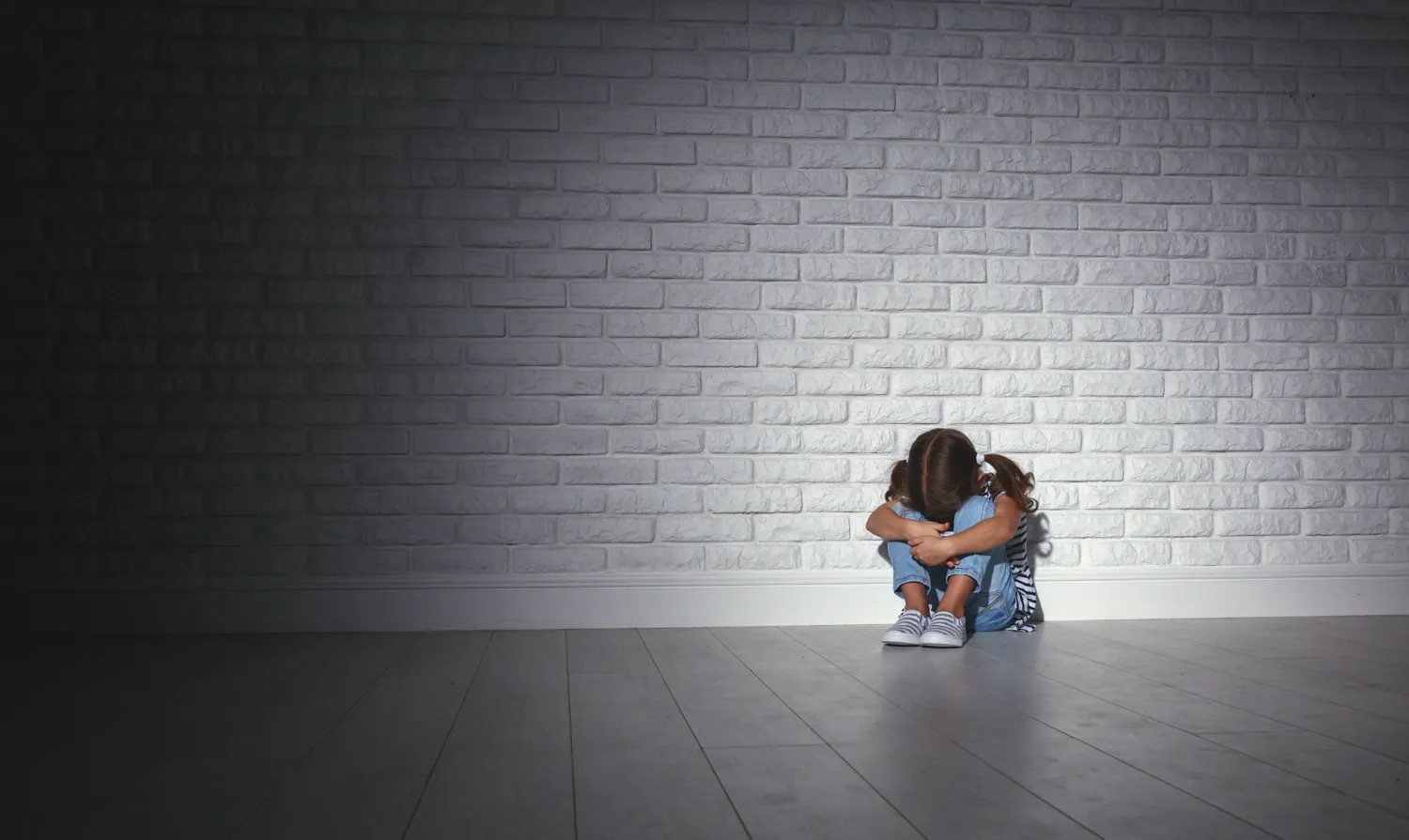 A child is sitting alone in a room, leaning against the wall and with her head burried in her knees.