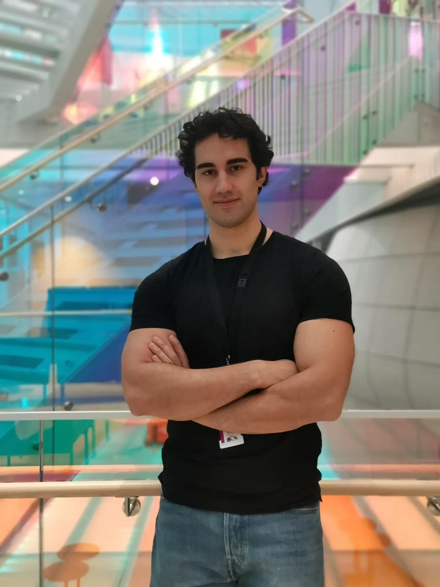 A man wearing a black T-shirt and jeans. He is standing with his arms crossed and is photographed indoors.