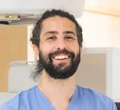 Portrait image of a man with dark hair and beard dressed in blue scrubs.