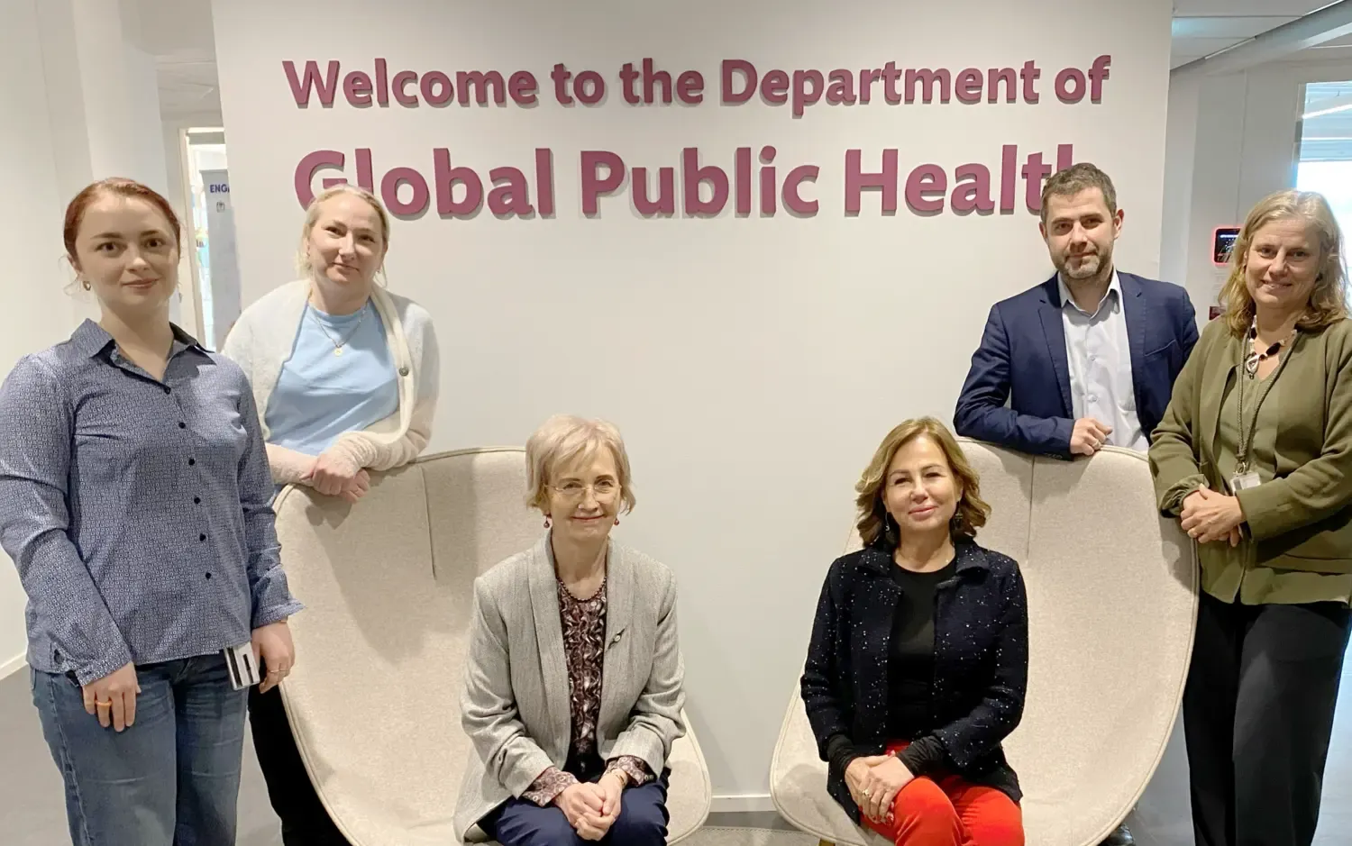 Three women and one man stan in front of a sign with the text Welcome to Department of Global Public Health. In front of them two women sit in armchairs.
