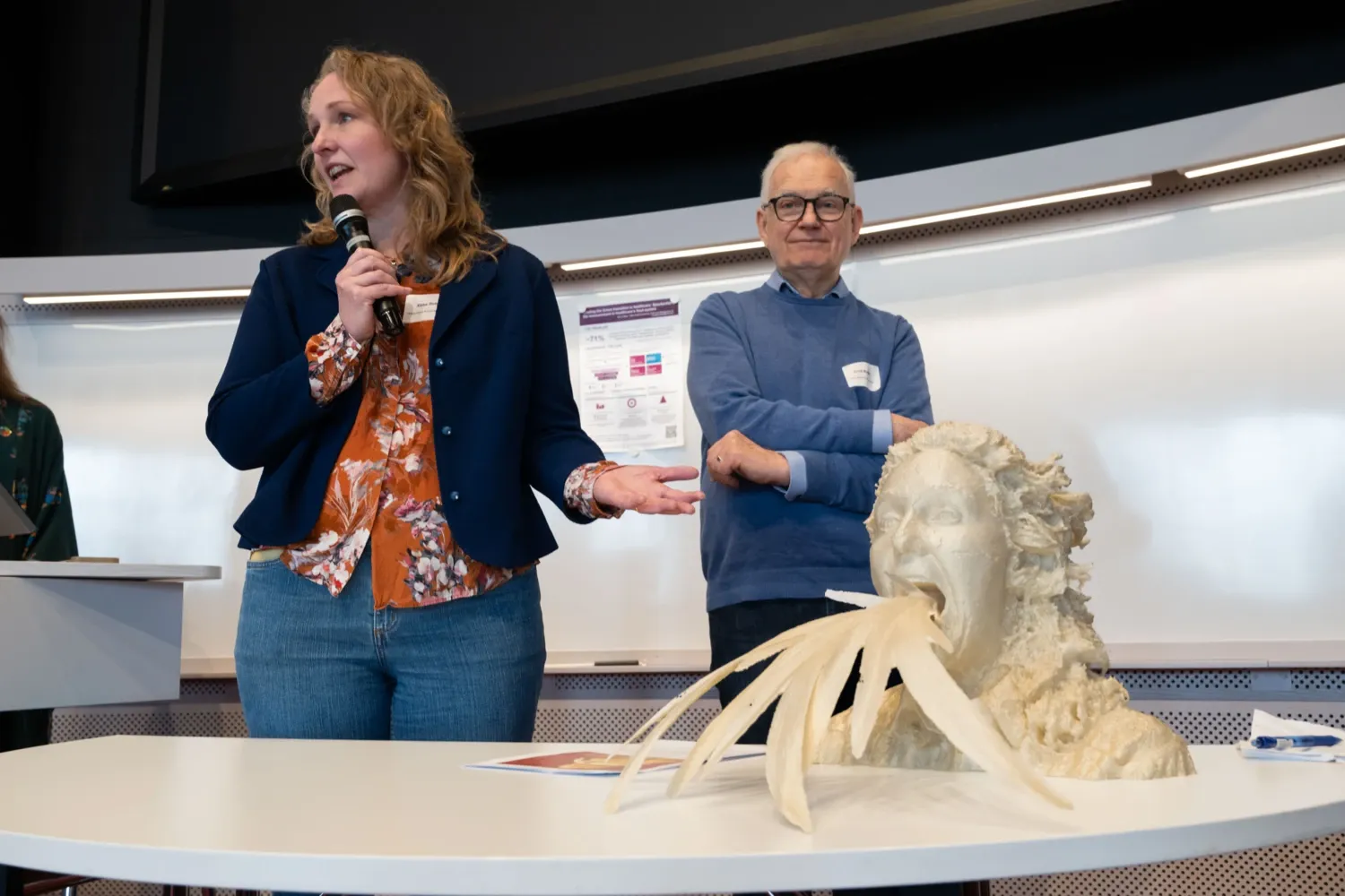 A three-dimensional artwork depicting a head with plant-like forms emerging from the mouth stands on a table in a lecture hall. In the background, participants at KI’s Sustainability Day can be seen.