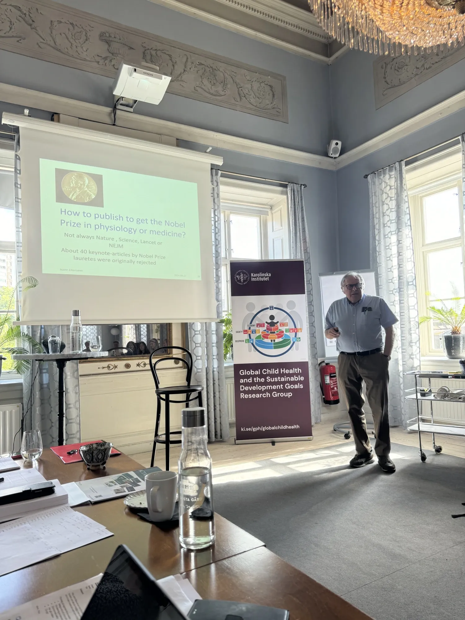 Hugo Lagercrantz next to a screen displaying a presentation. A banner beside him reads, "Global Child Health and the Sustainable Development Goals Research Group." The room includes a table with various items.