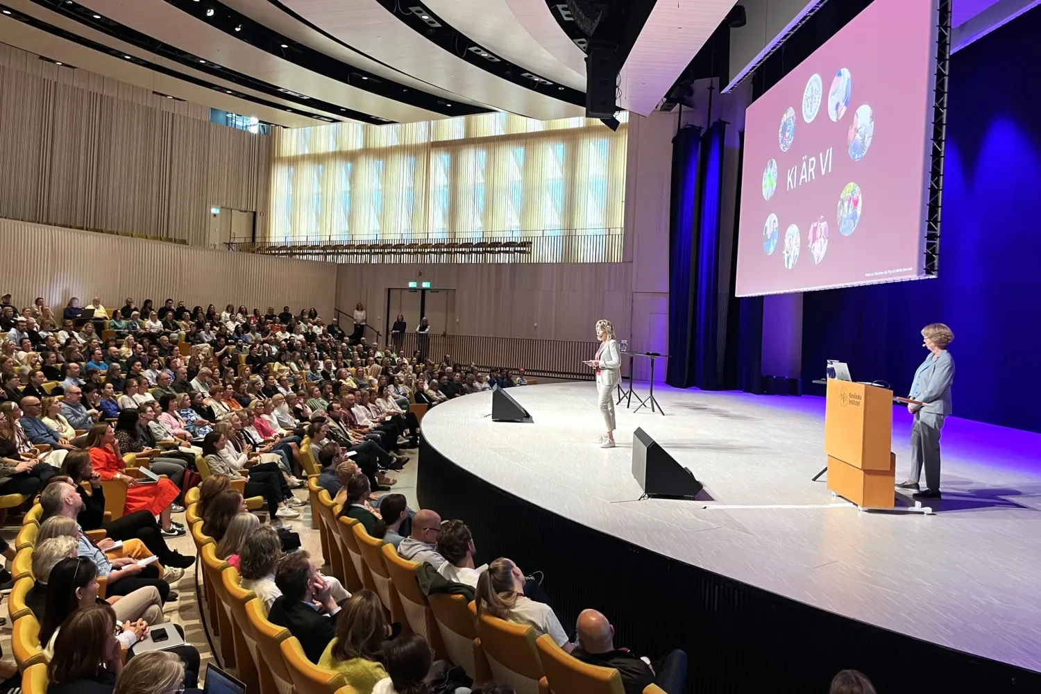 The President, dressed in a beige suit, stands on the stage in Aula Medica and speaks to a large audience in yellow seats in front of her. In the background, there is a picture with the text “KI is us.”