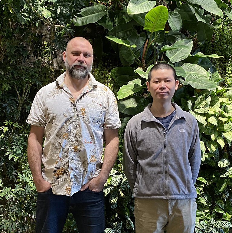 Two men standing infront of a wall of green plants.