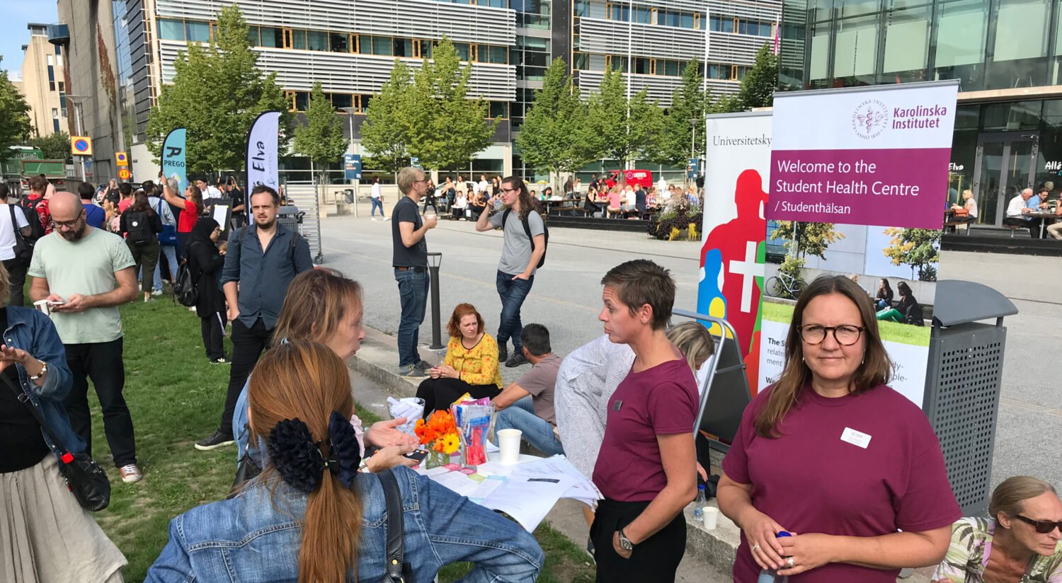Photo of a female staff member from KI's Student Health Services wearing glasses and a KI purple profile t-shirt, looking at the camera, surrounded by visitors at Campus Day at the Flemingsberg campus.
