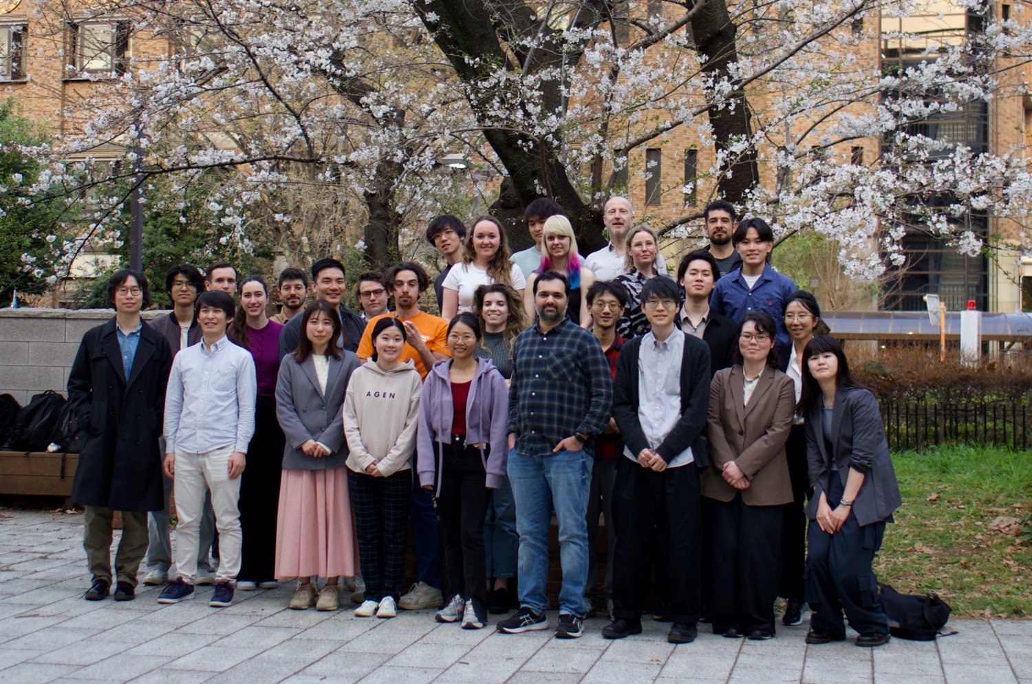 A group of people standing and kneeling in front of a blooming cherry blossom tree. The background includes a large building with multiple windows and some greenery.