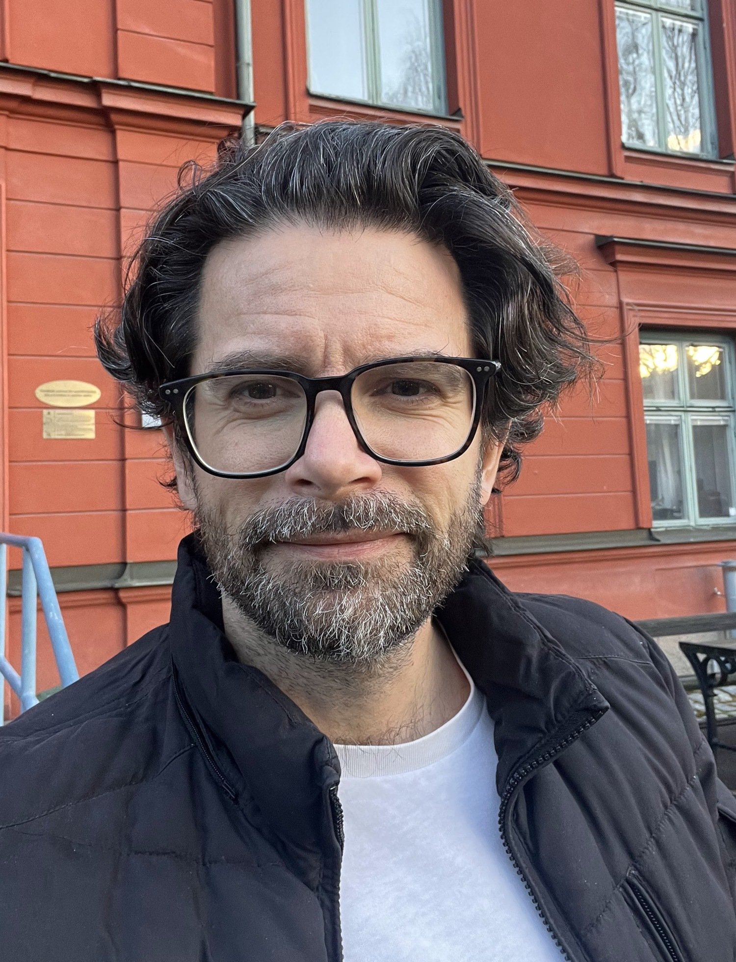 Portrait of Gergö Hadlaczky. He is standing outdoors in front of a red building. He is wearing glasses, a white tshirt and a black jacket.