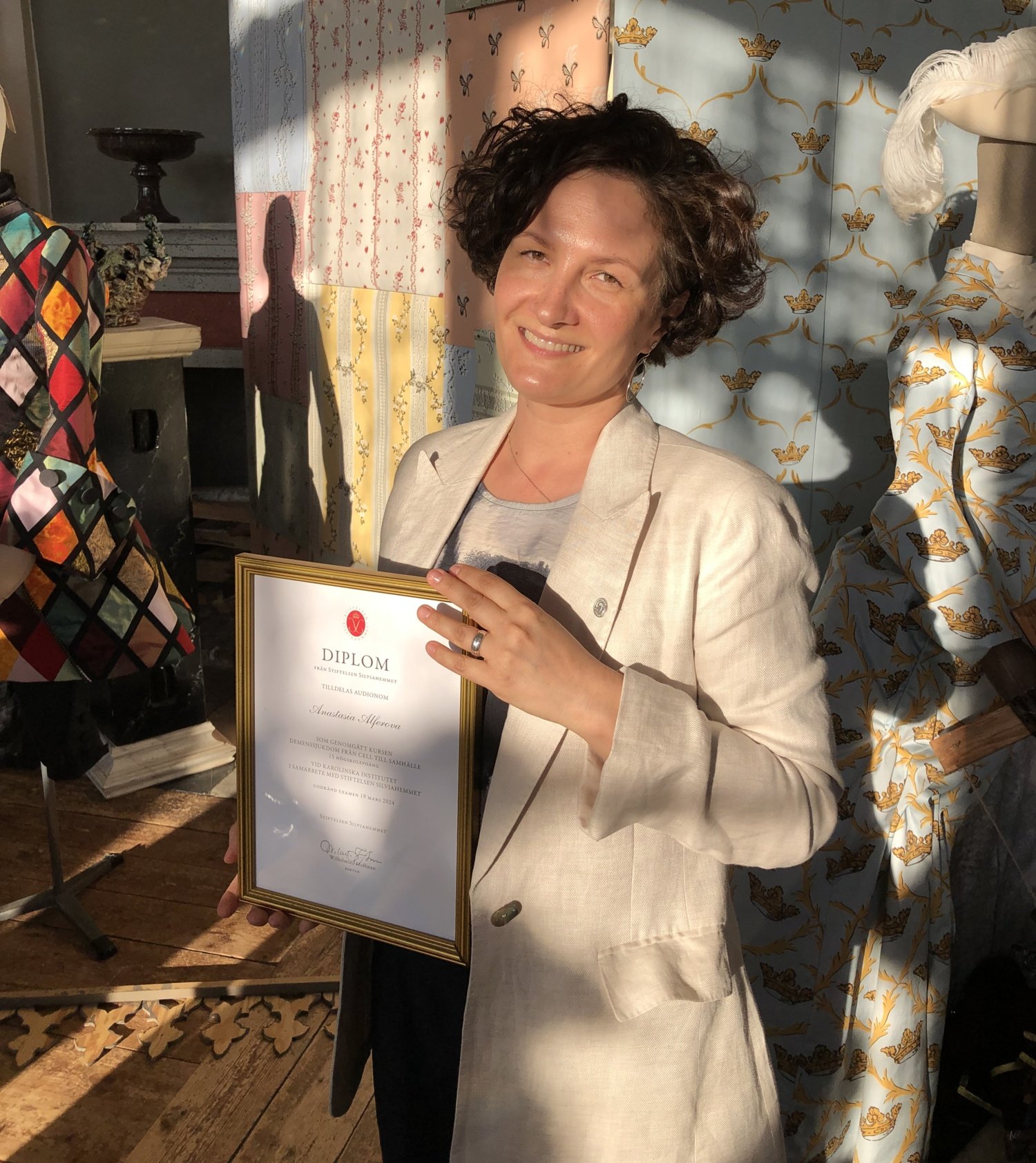 A photograph of a woman in afternoon light bright blazer and curly brown hair holding a framed diploma.