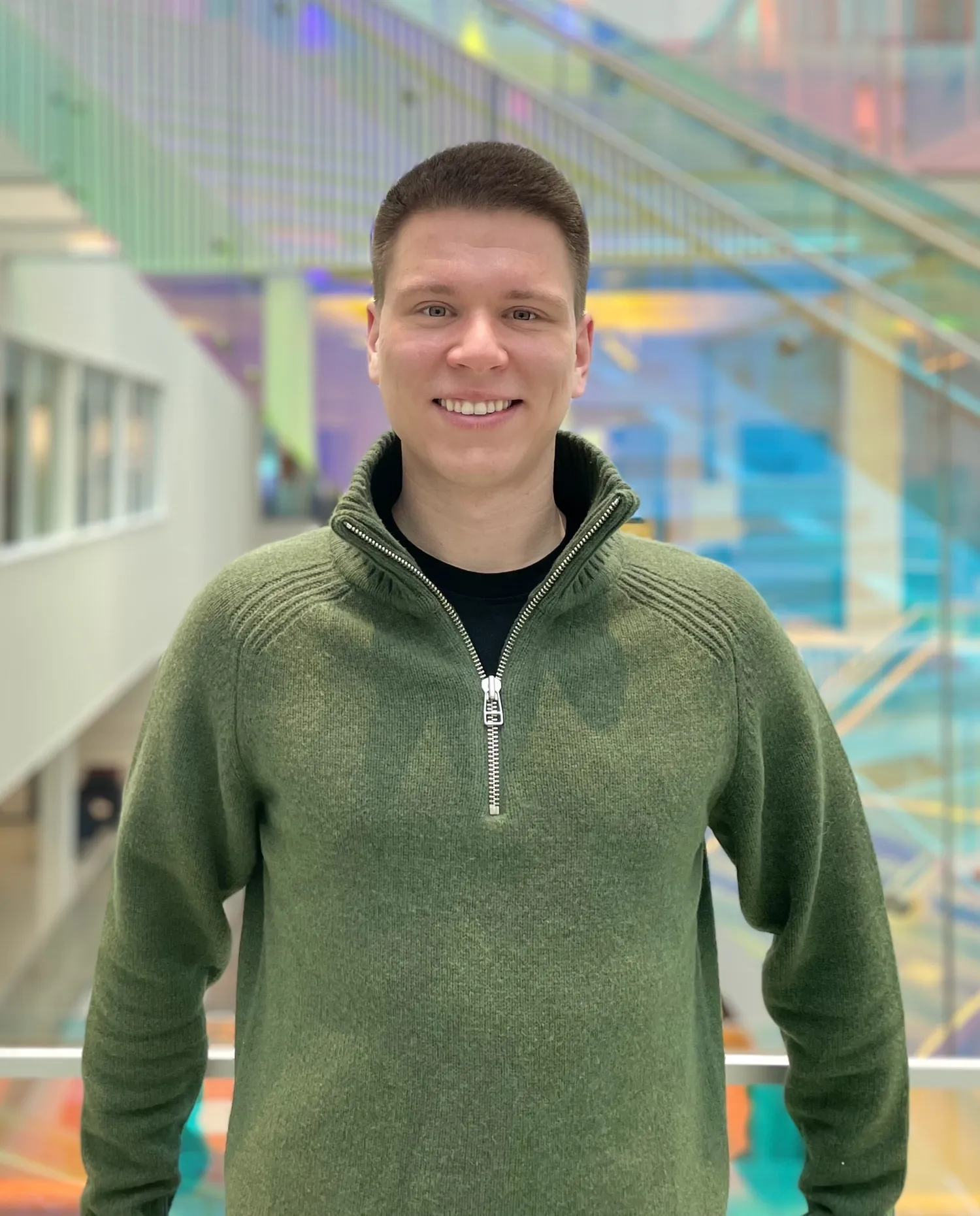 A smiling man with dark hair wearing a green jumper. Photographed indoors.