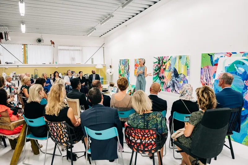 Group pf people listening to woman speaking, standing in front of paintings.