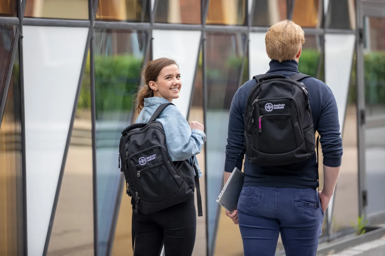 Two students outside Aula Medica walking away with their KI-backpacks. One turns around and smiles at the camera. || Two students walk in front of Aula Medica and have KI backpacks on their backs.