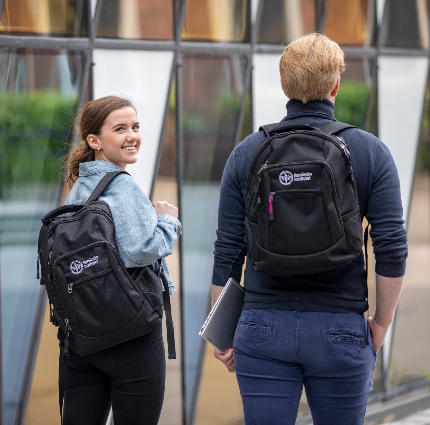Two students outside Aula Medica walking away with their KI-backpacks. One turns around and smiles at the camera. || Two students walk in front of Aula Medica and have KI backpacks on their backs.