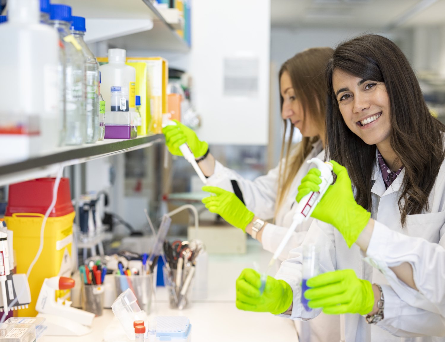 Three femare researchers standing with test tubes and pippettes in lab environment. One of them is looking in to the camera.