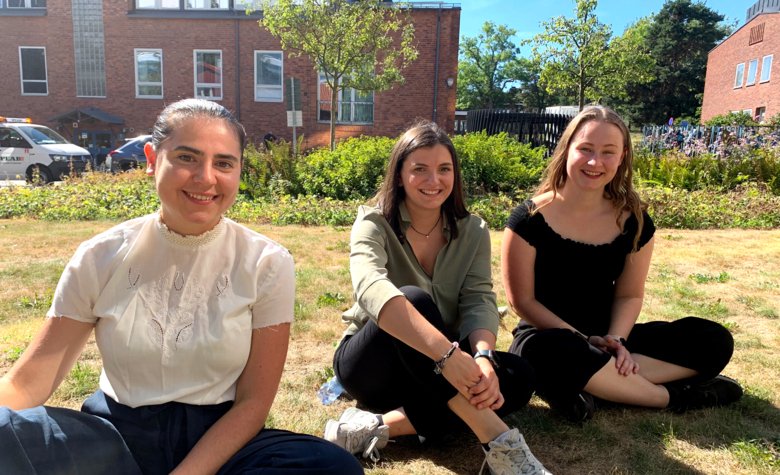 Three students sit outside Aula Medica on the grass in sunny weather.