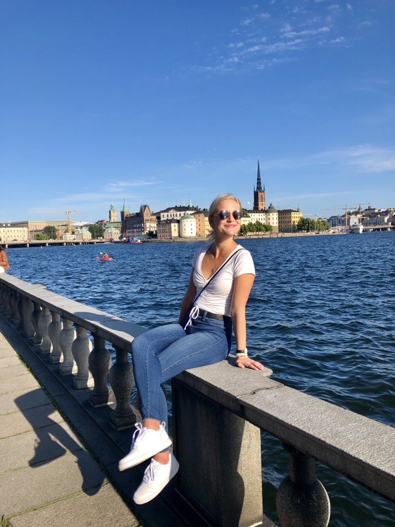 Female student sitting in front of the water. In the background you can see Stockholm in the sun.