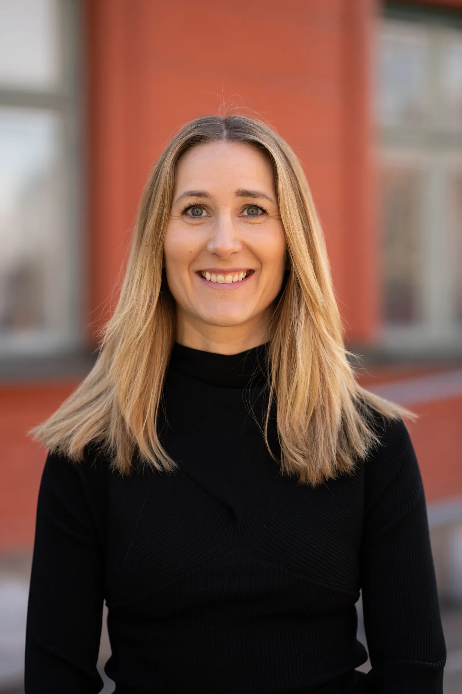 Photo depicting a woman with light-colored hair and a black sweater. She is standing outdoors in front of a brick-red building.
