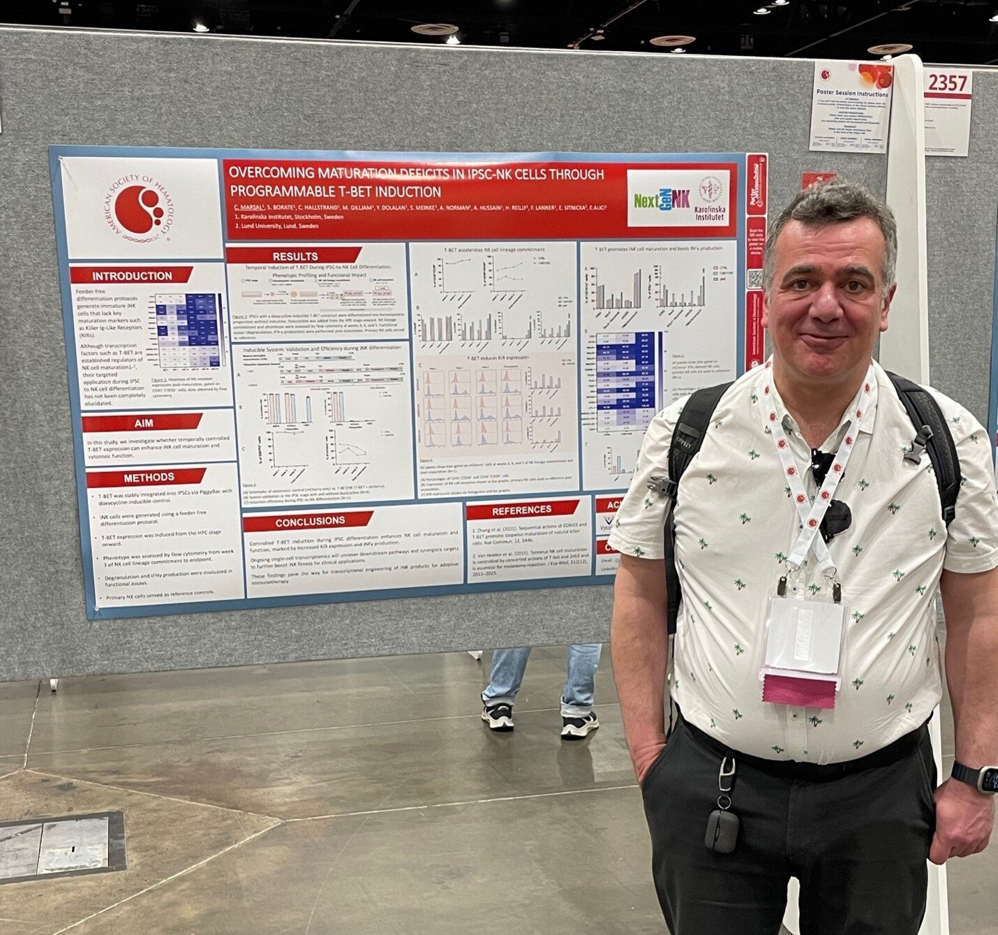 A man wearing a light-coloured short-sleeved shirt and dark trousers stands in front of a notice board displaying a scientific poster.