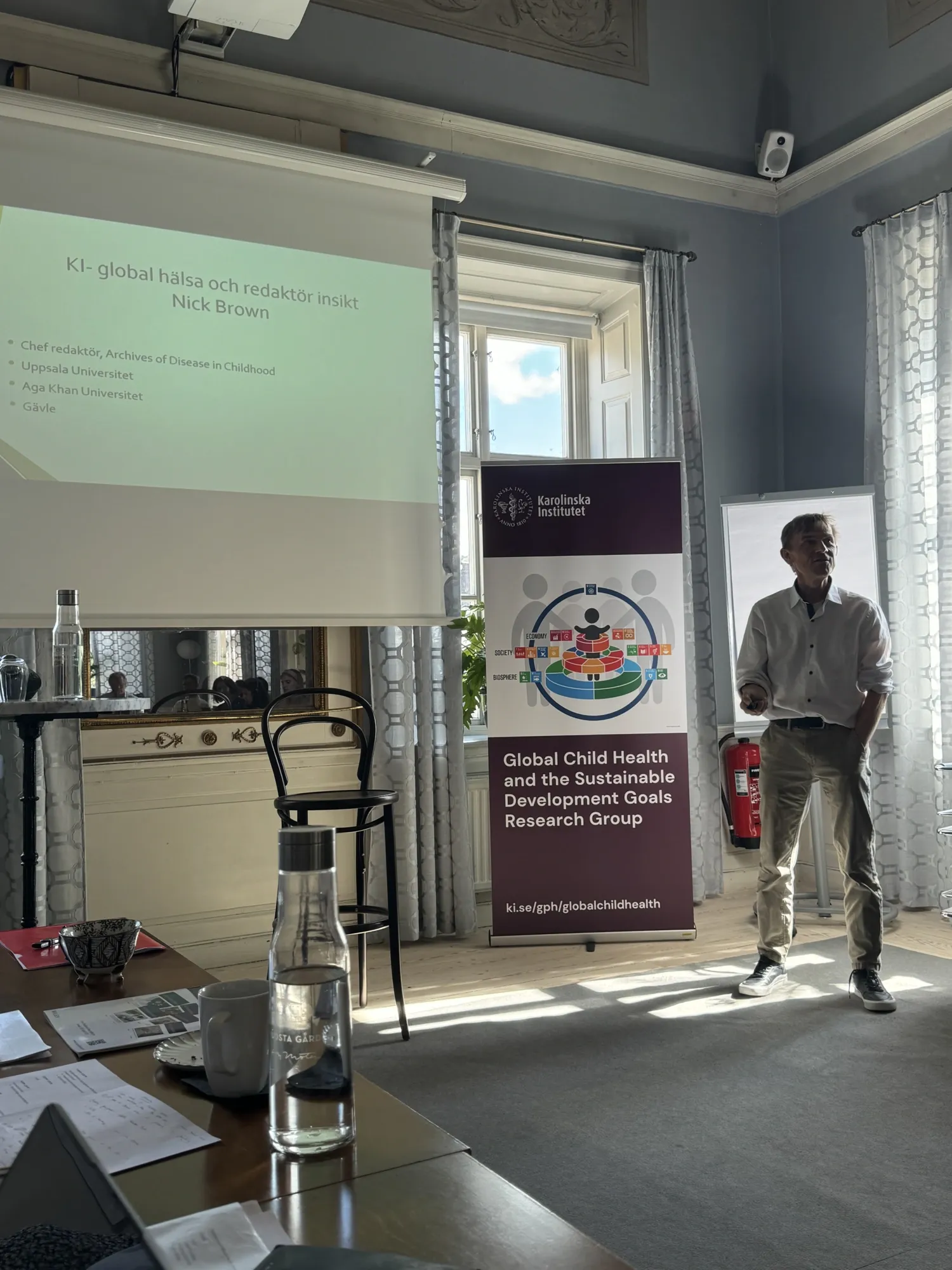 Nick Brown next to a screen displaying a presentation. A banner beside him reads, "Global Child Health and the Sustainable Development Goals Research Group." The room includes a table with various items.