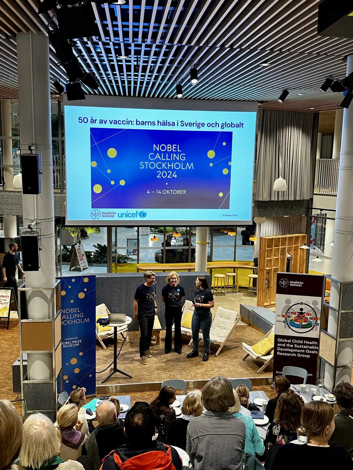 A group of three speakers stands on a stage in front of an audience, with a large screen displaying "NOBEL CALLING STOCKHOLM 2024" and event details. The setting is a modern venue with wooden accents and seating arranged for attendees. The speakers are di