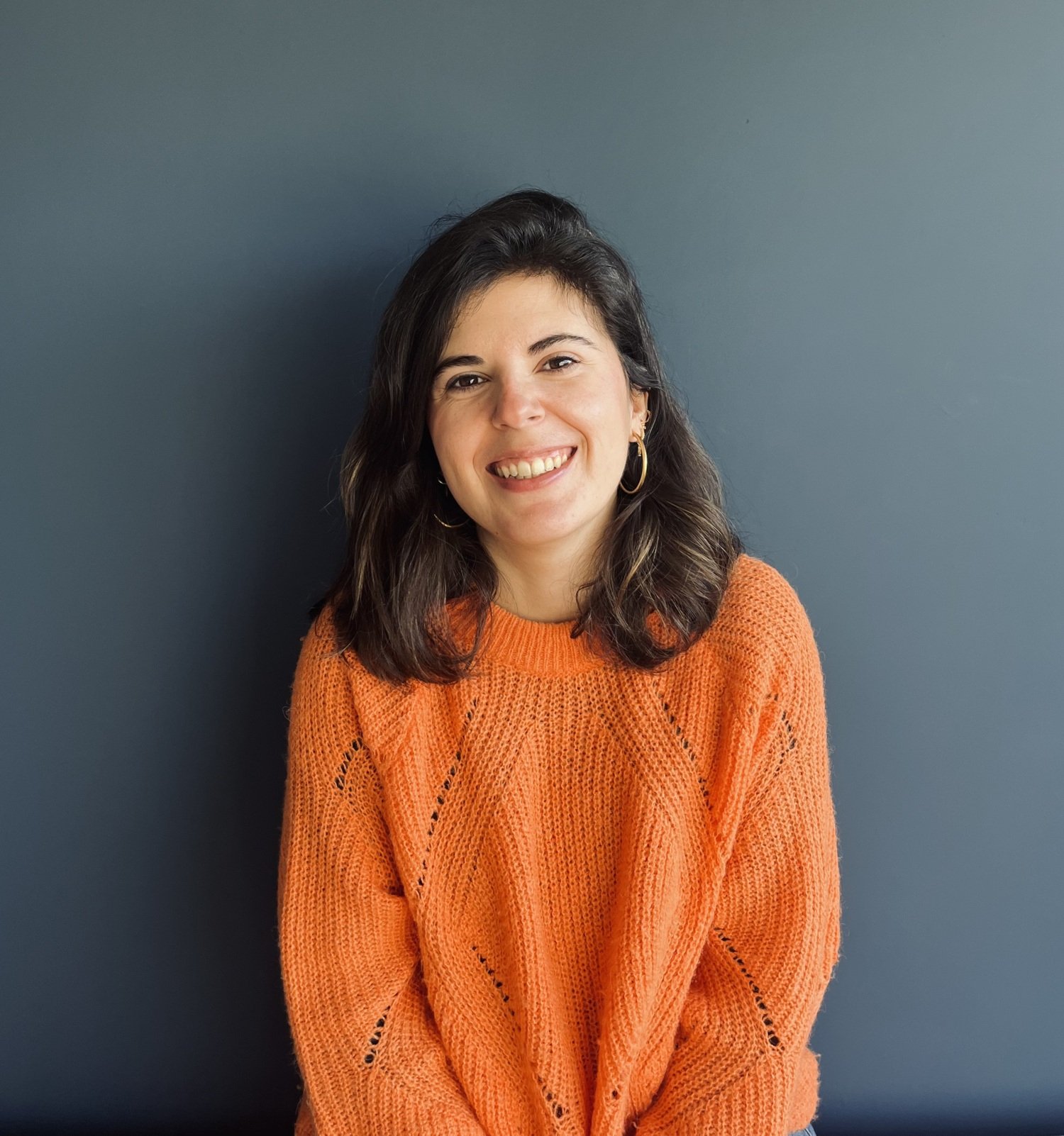 Portrait of Olga Rivera Ballesteros, who has dark hair and is wearing an orange jumper. She is smiling big and is sitting in front of a dark blue wall.