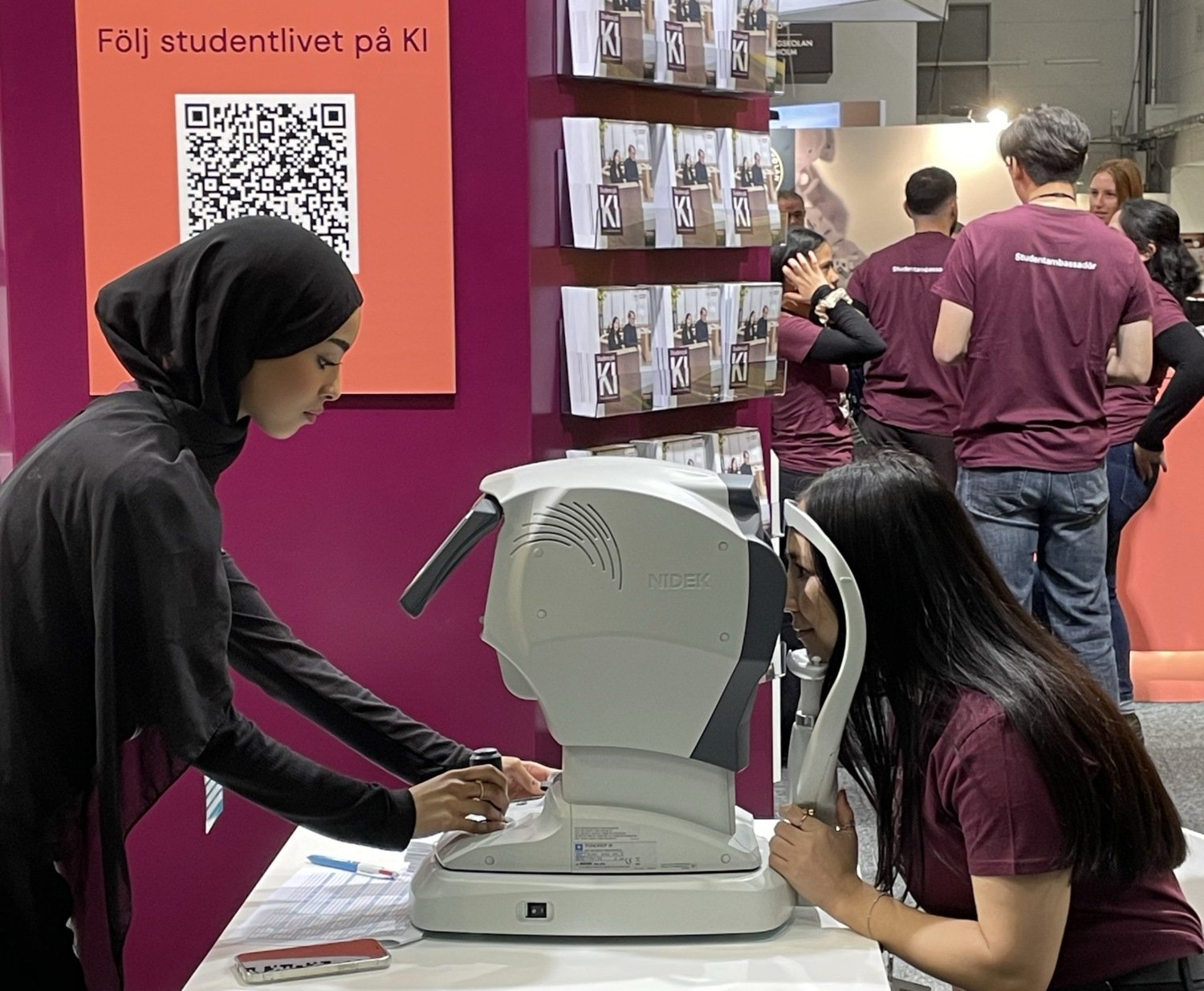 A KI student stands on one side of an eye examination machine while another student looks into it, at the Saco student fair.