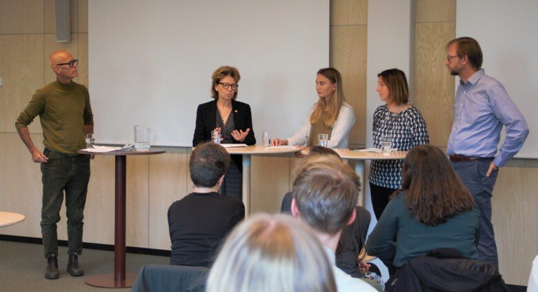 Five people, two men and three women, standing by two tall tables. They are the panel at a seminar