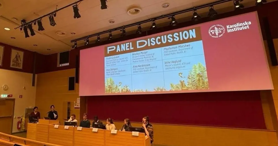Eight persons sitting in a panel under a large screen with the text Panel Discussion.