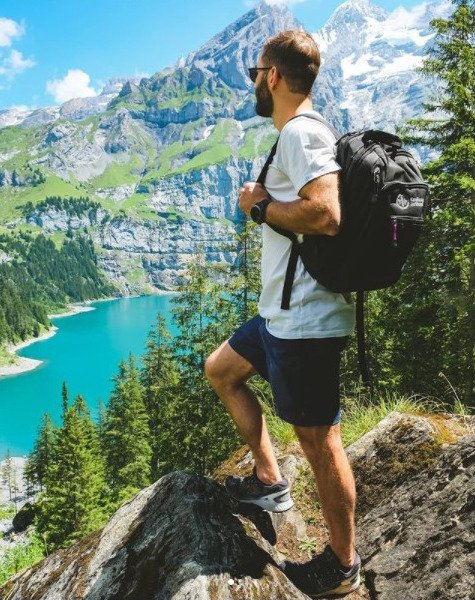 A student is standing with his back to the camera, wearing a backpack. He is standing on a cliff and looking at the water and mountains.