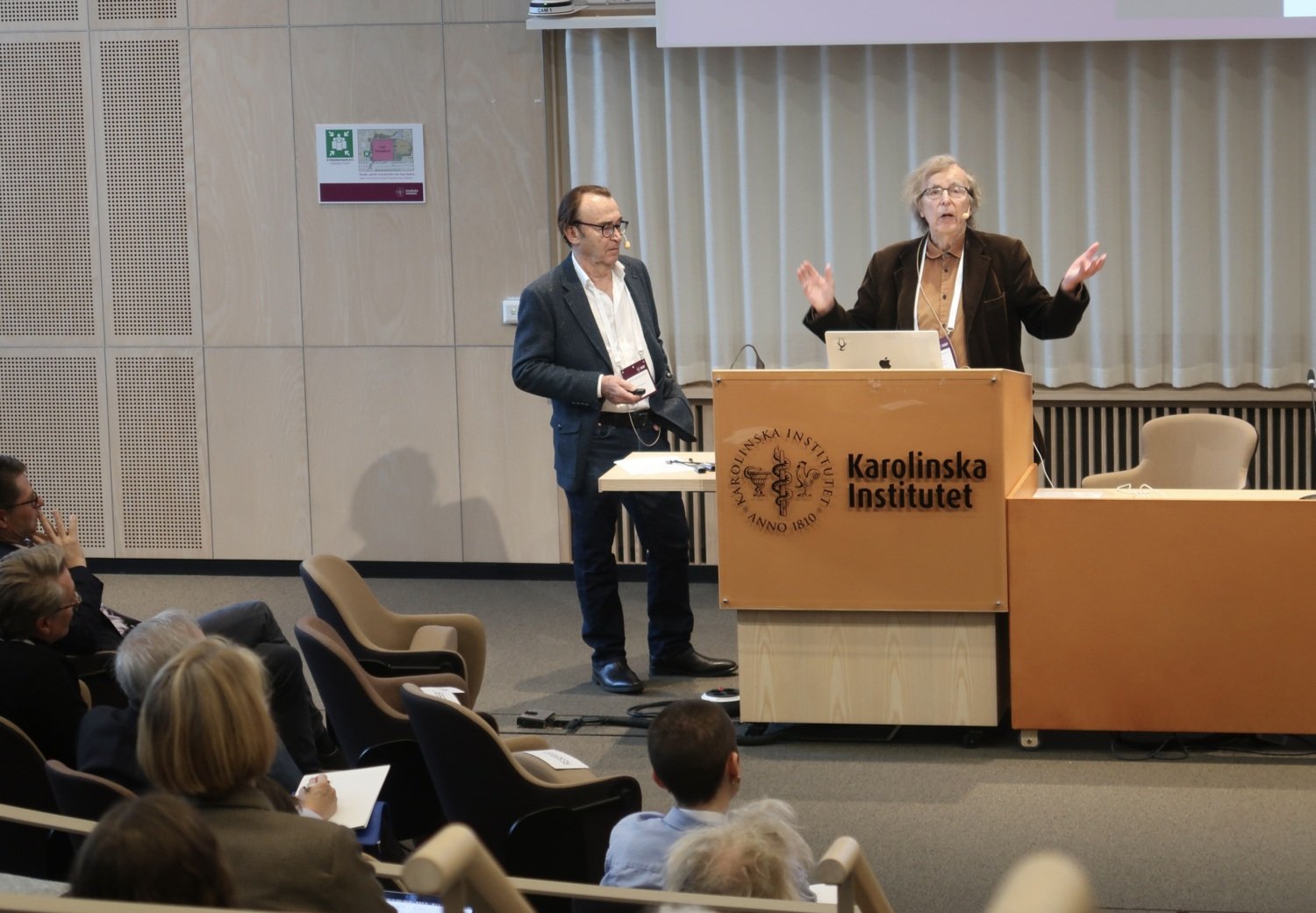 Two men are standing at the lectern at the front of a lecture hall and speaking.
