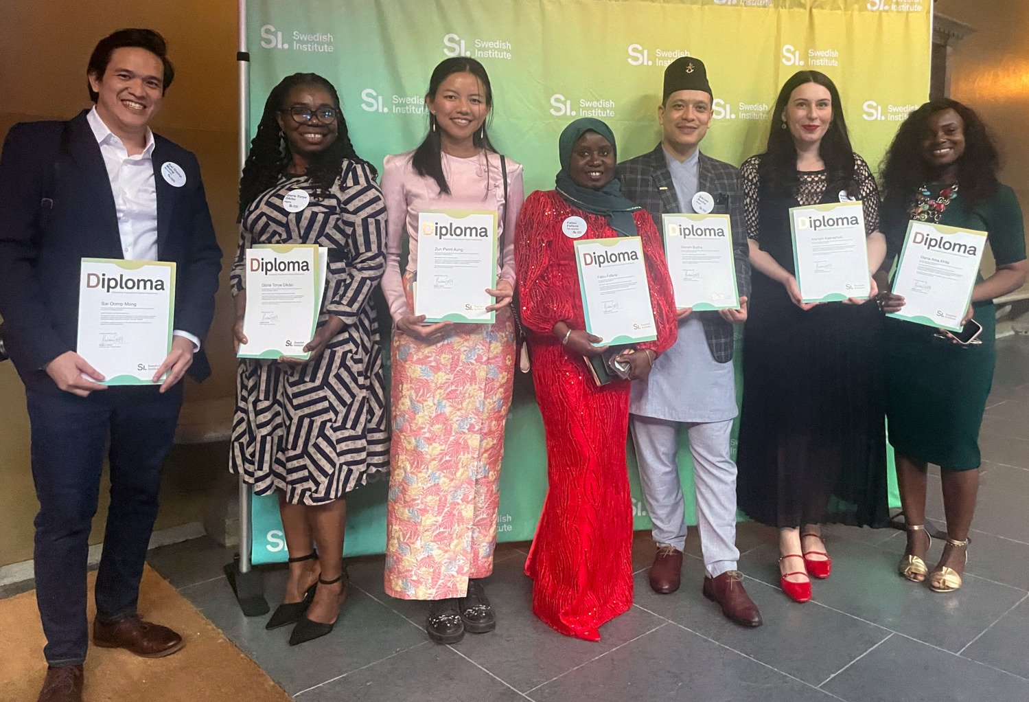 Seven international students smiles and show their diplomas.