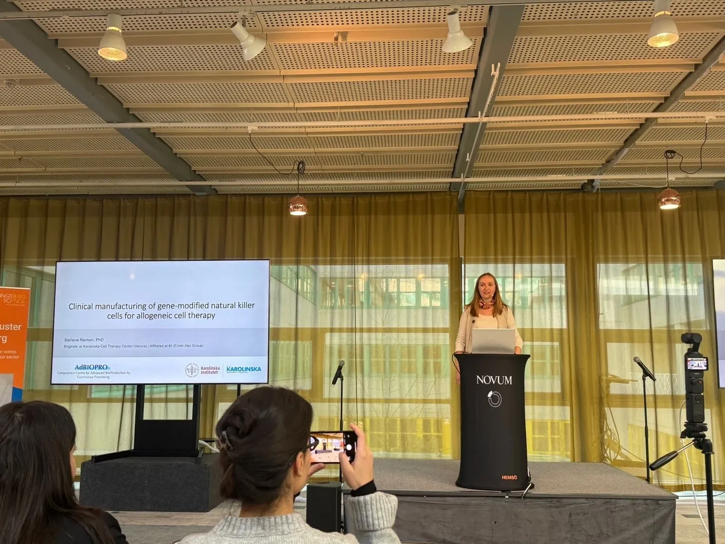 A woman is standing behind a lectern on a stage, giving a lecture. Next to her is a projection screen showing a presentation.