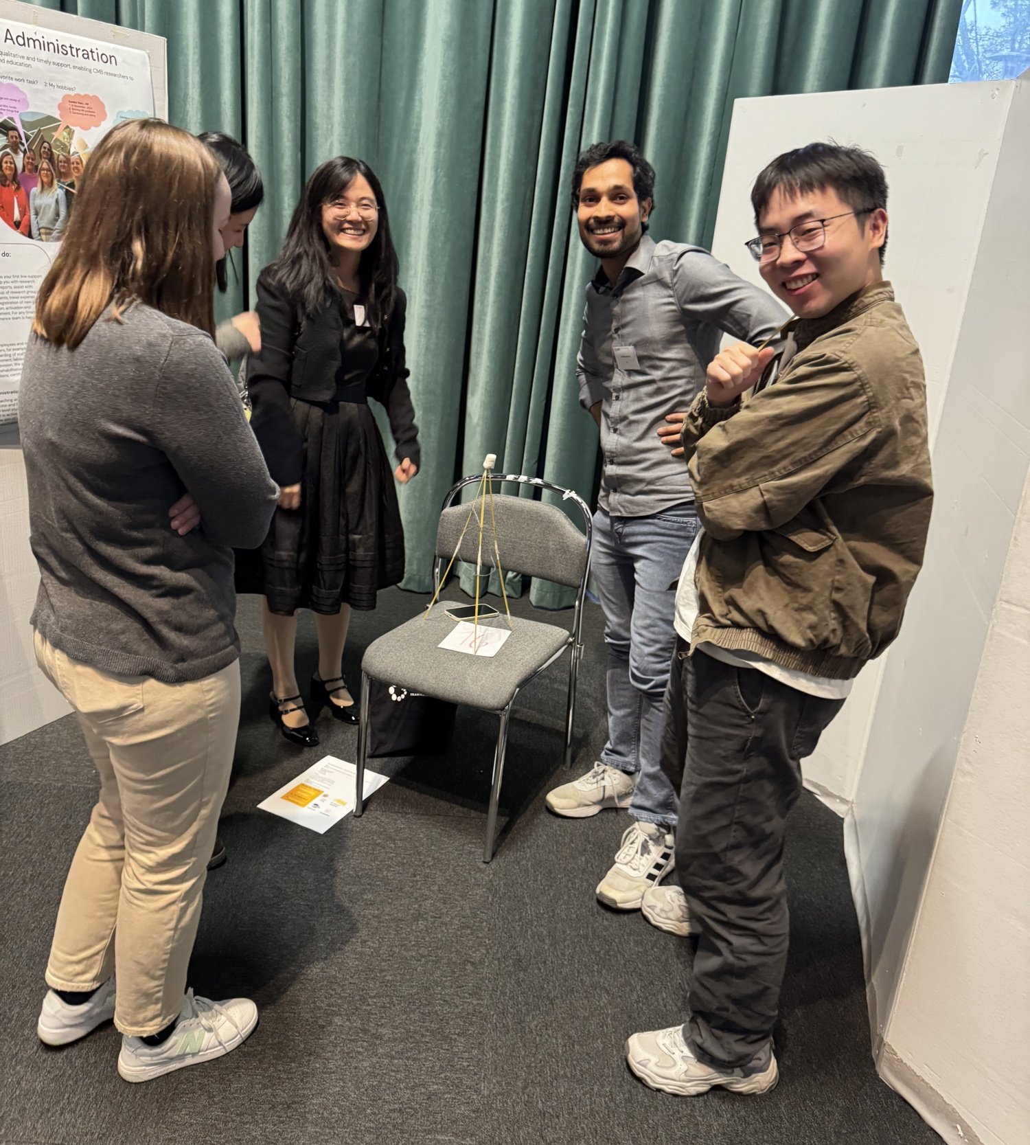 Five people standing around a chair, on the chair is a tower build of spagetti.